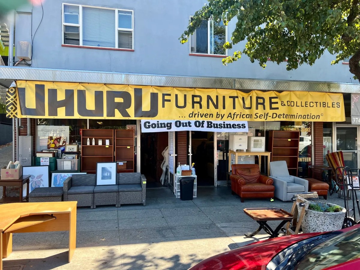 A large yellow banner hanging over a storefront reads, "Uhuru Furniture and Collectibles ... driven by African Self-Determination." Another banner below the yellow one reads, "Going Out of Business." Furniture and housewares line the sidewalk outside of the store.