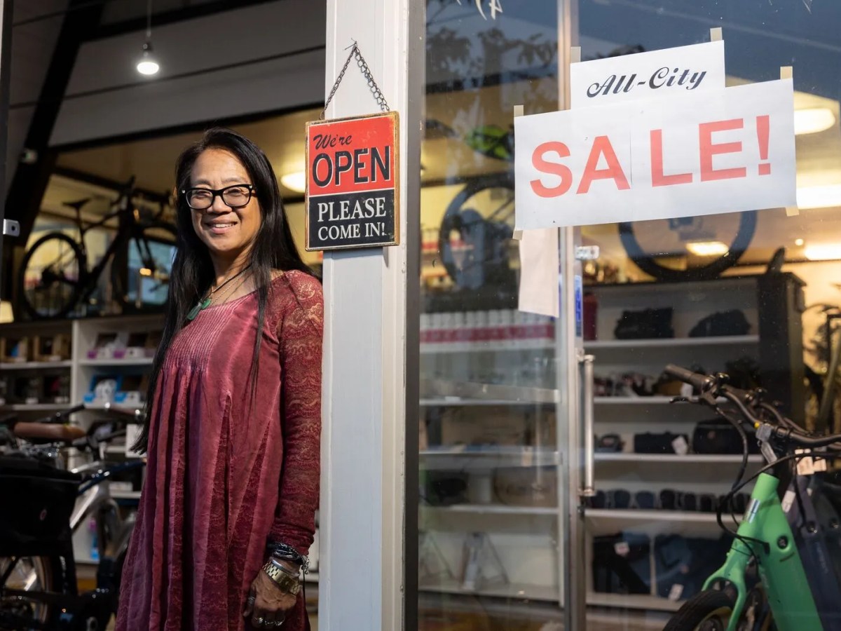 A woman wearing glasses and a burgundy dress leans on one side of an entryway. A sign on the entryway reads, "We're open, please come in!" A large glass window on the right has another sign reading, "All City sale!"