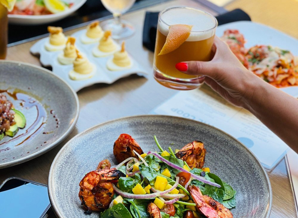 A hand holds a drink above a table with a variety of plated food on it.