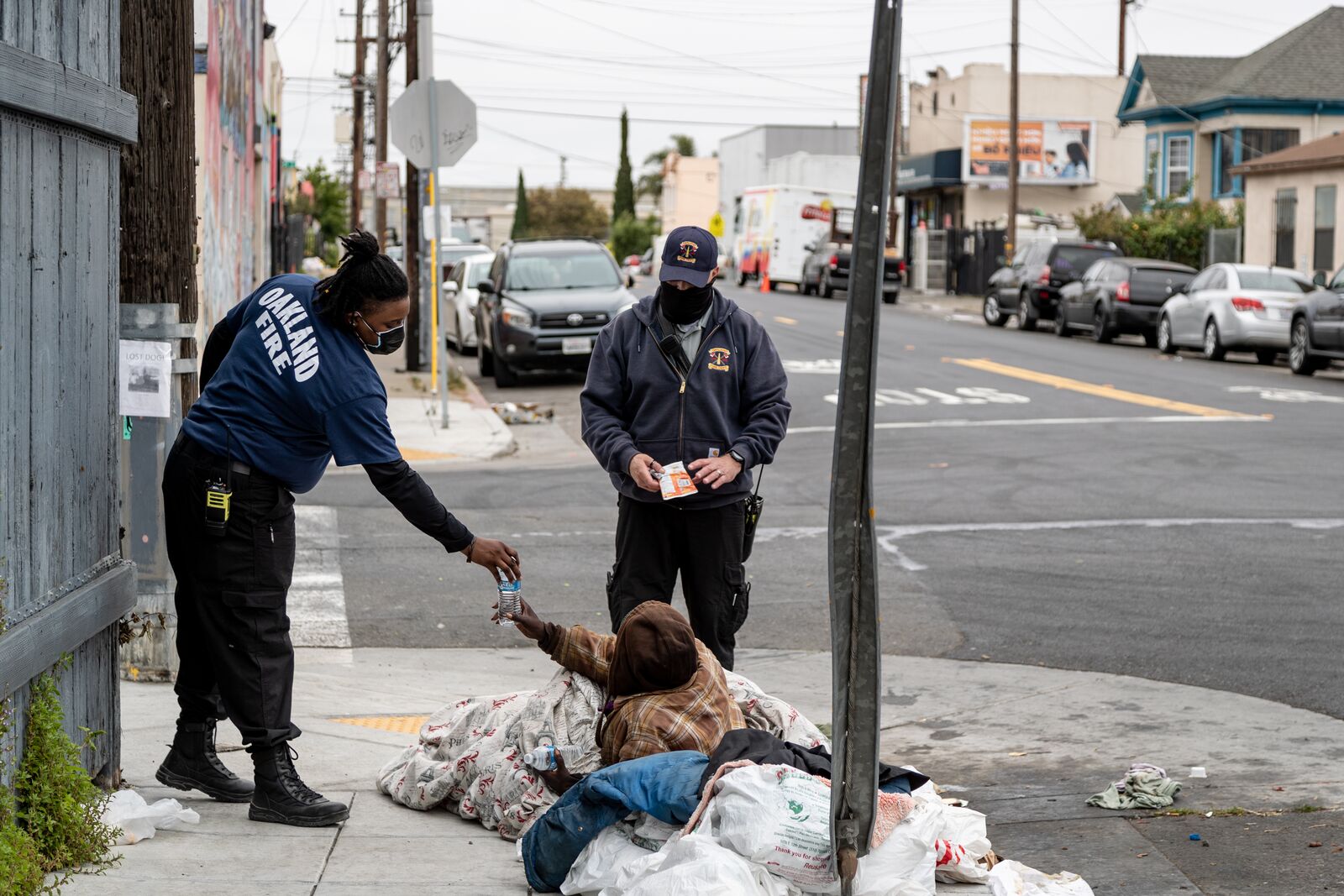 Two people wearing fire department shirts and wearing masks giving a water bottle to a man lying on the street wrapped in a blanket.