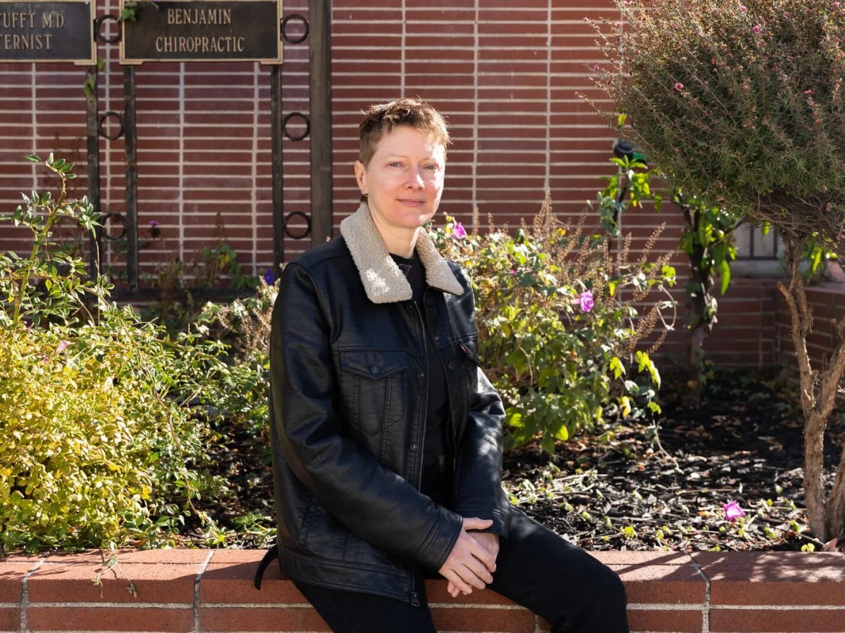 A woman in a brown leather jacket sits on a brick ledge outside of a small medical building.