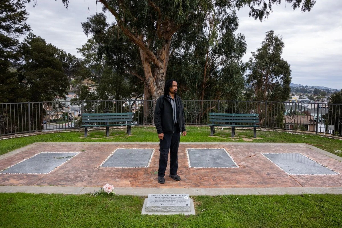 A Black man stands on a brick and concrete slab in front of four large plaques in the middle of a lawned and tree-lined cemetery on a hill, with homes in the background.