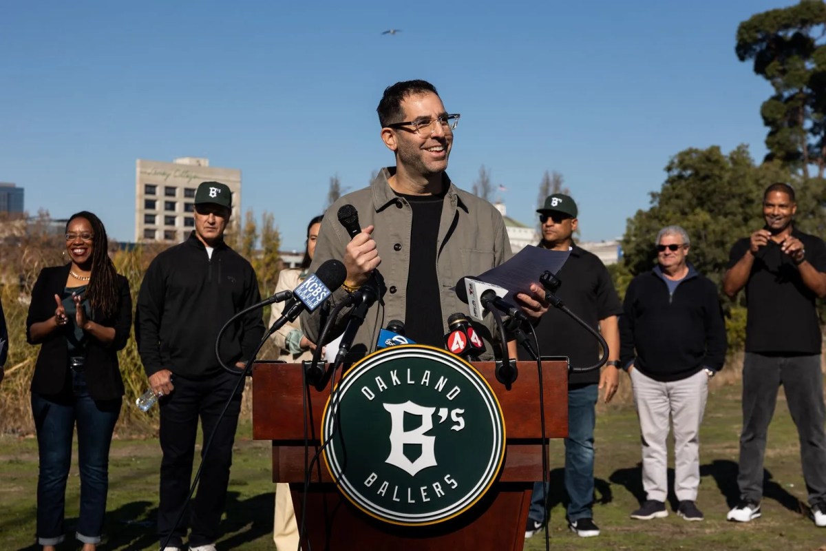 A man speaks outdoors at a podium. A baseball team logo, a prominent "B" for Ballers is on the podium. Behind him are Oakland Mayor Sheng Thao and other officials.