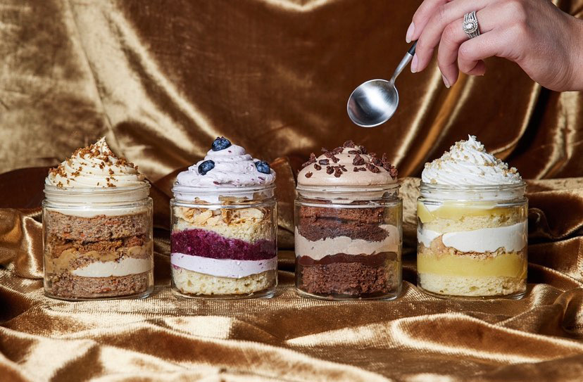 Four mini cakes from Full Belly Bakery sit in a row and a hand holding a spoon hovers above them,