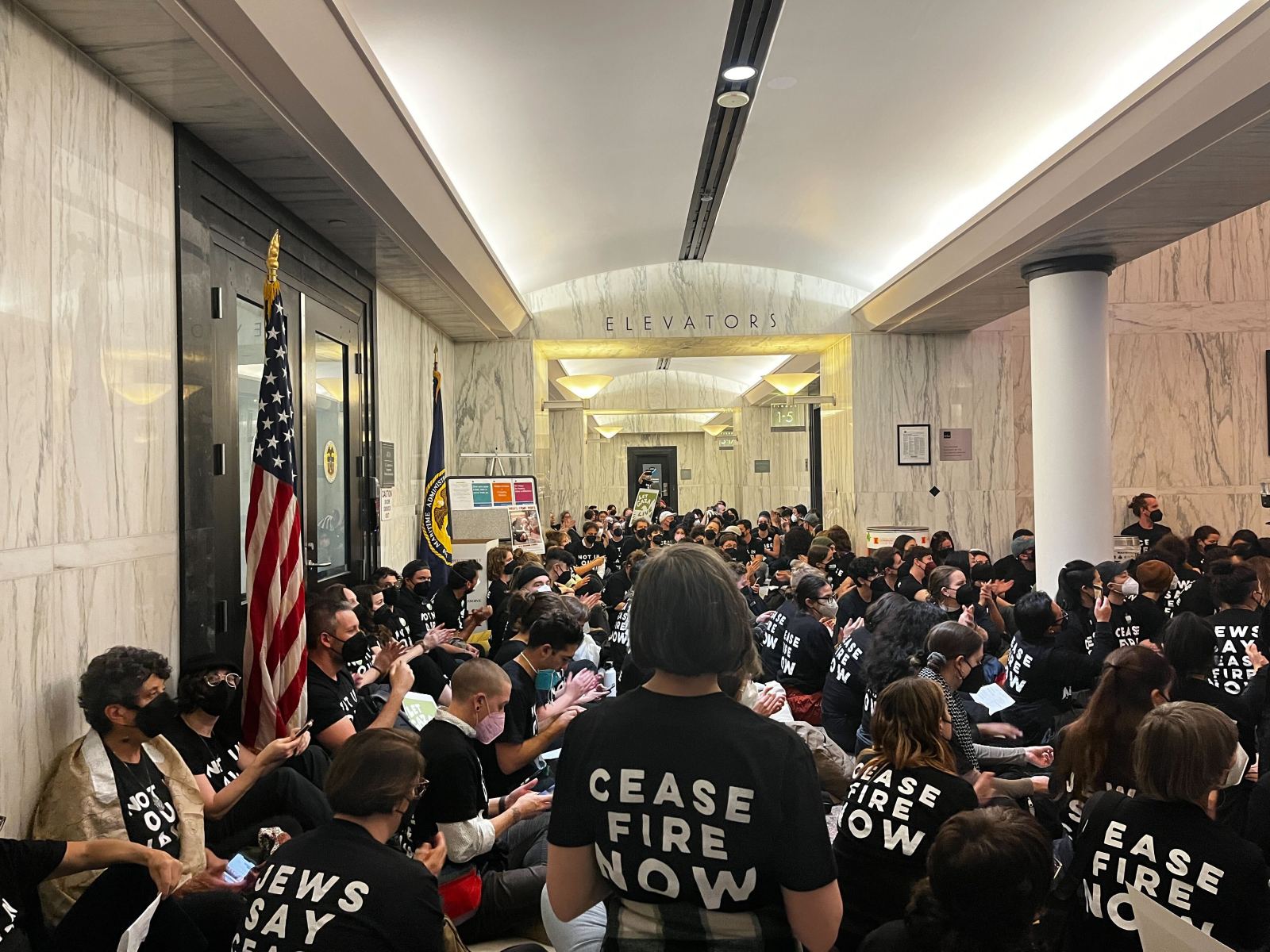 Scores of people in black shirts, some of which say "Ceasefire Now," sit inside the lobby of one of the towers of the Oakland Federal building.
