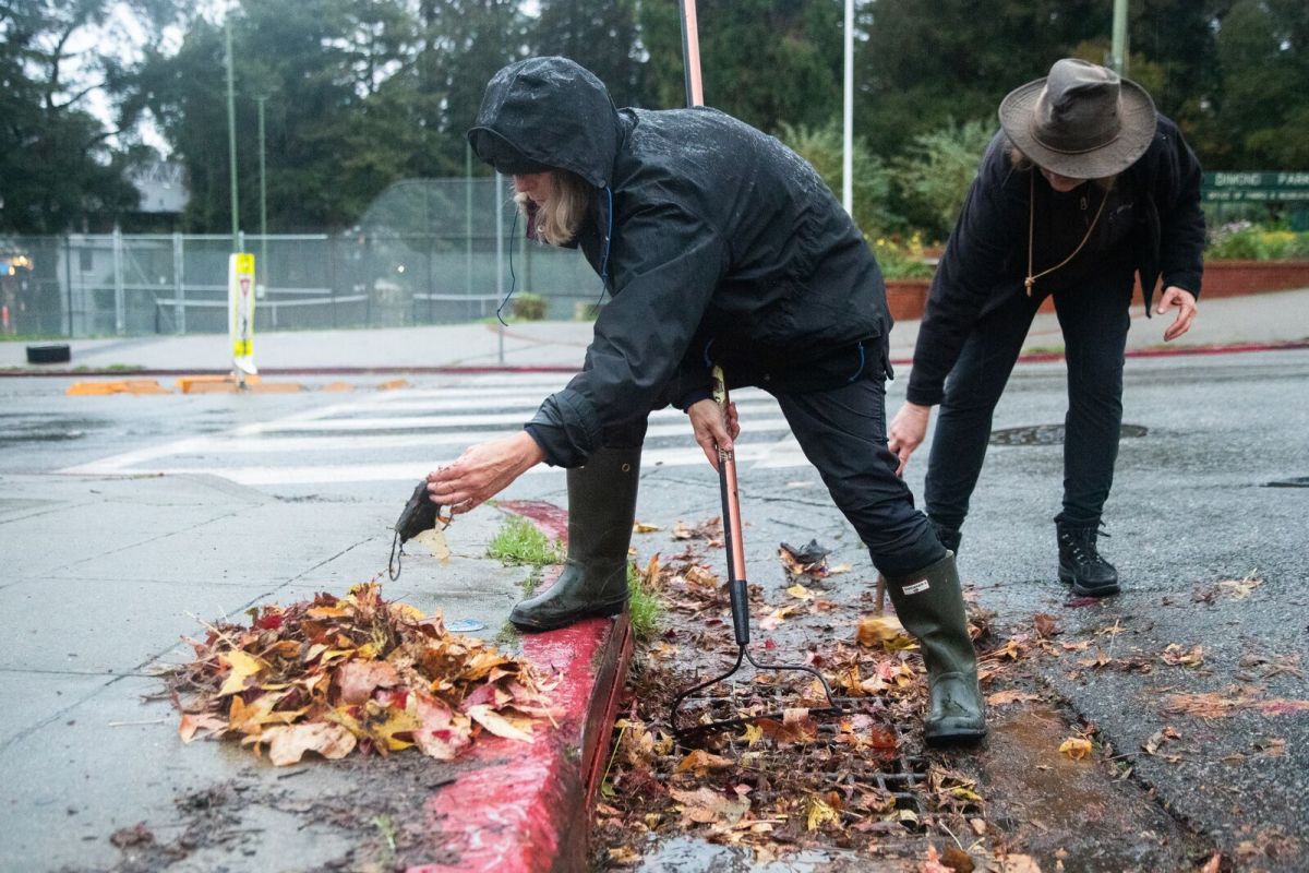 Two people dressed in black rain gear clearing a street gutter of fallen leaves