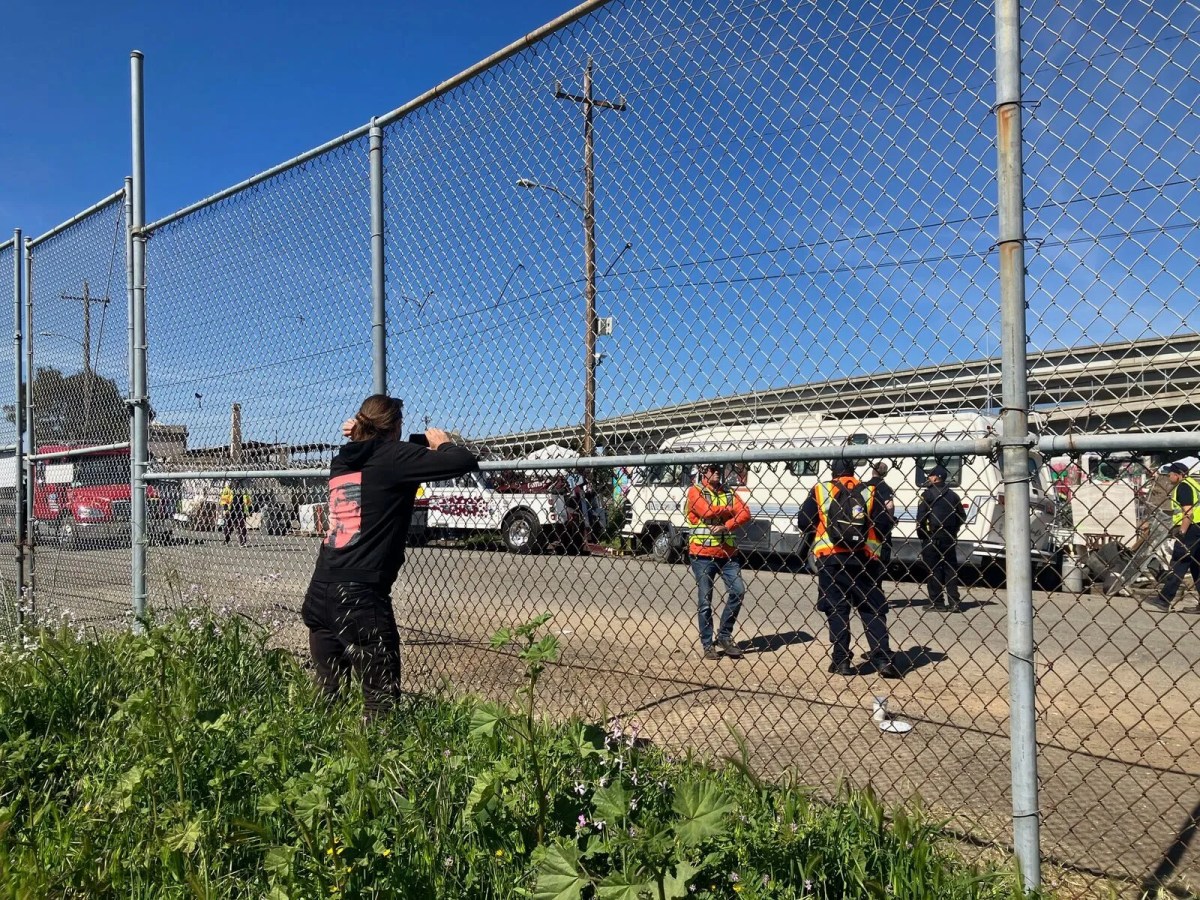 Person leaning against a large fence. Behind it are people in neon vests and RVs.