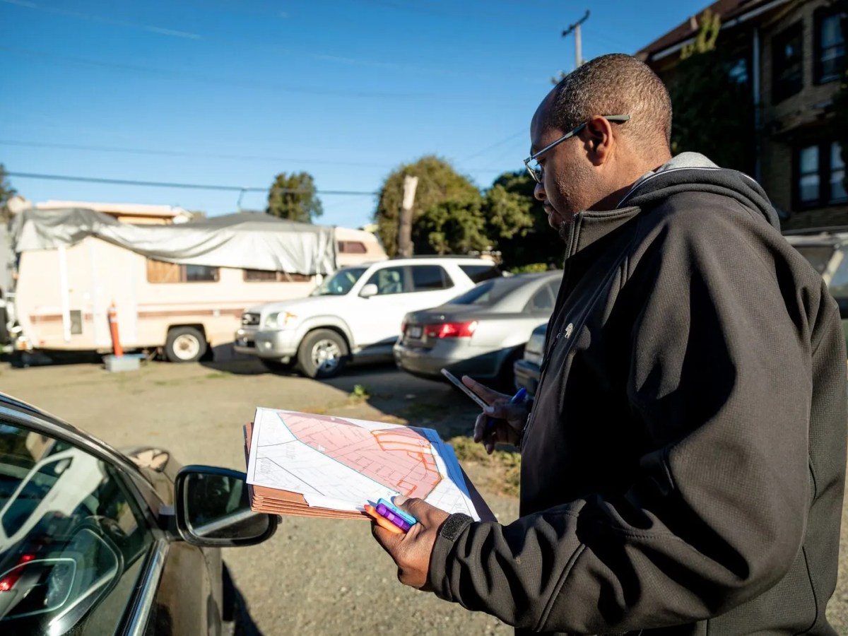 Loren Taylor stands outside of his car, holding a paper map of Oakland, looking at an RV in the distance.