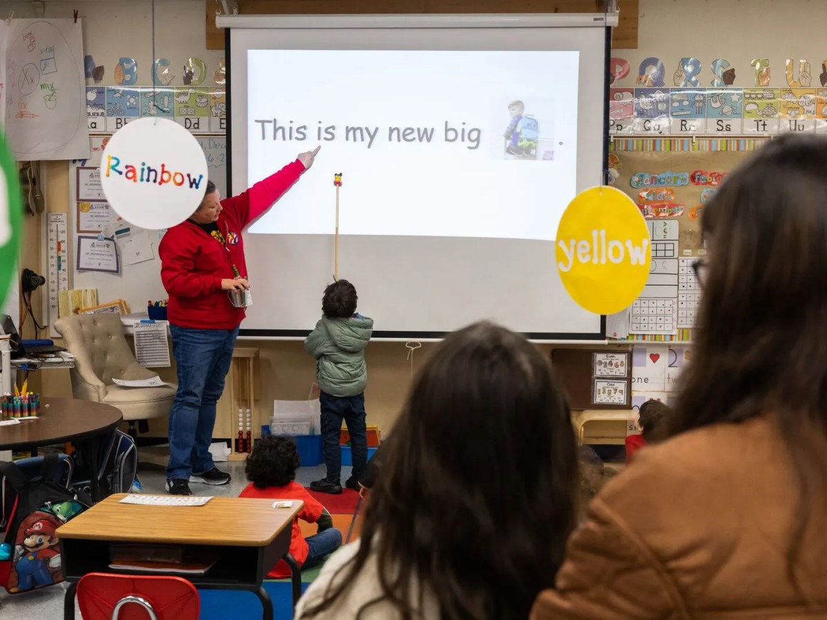 In a kindergarten classroom, a student uses a pointer to read the sentence "This is my new big backpack" shown on the projector screen while his teacher points with her finger.