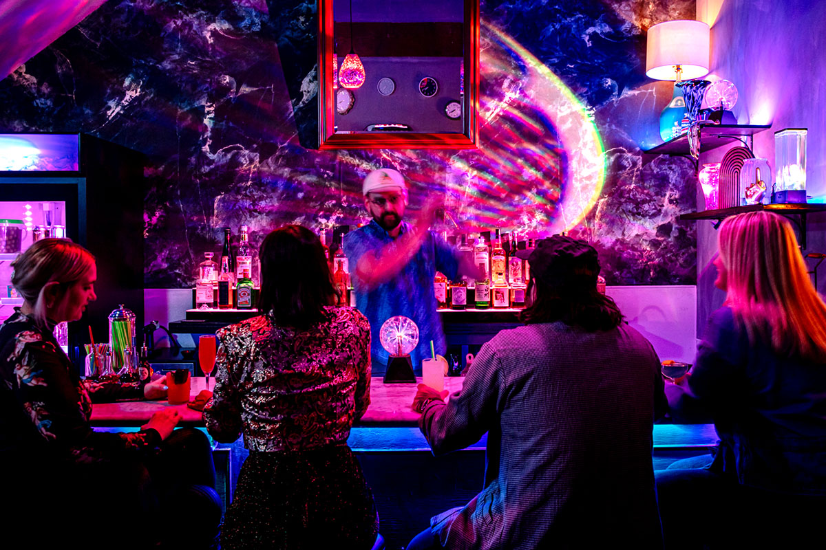 Ninth Life drink program director and co-owner Daniel Paez stands behind a dimly lit bar making a drink with a shaker as customers sit at the bar.