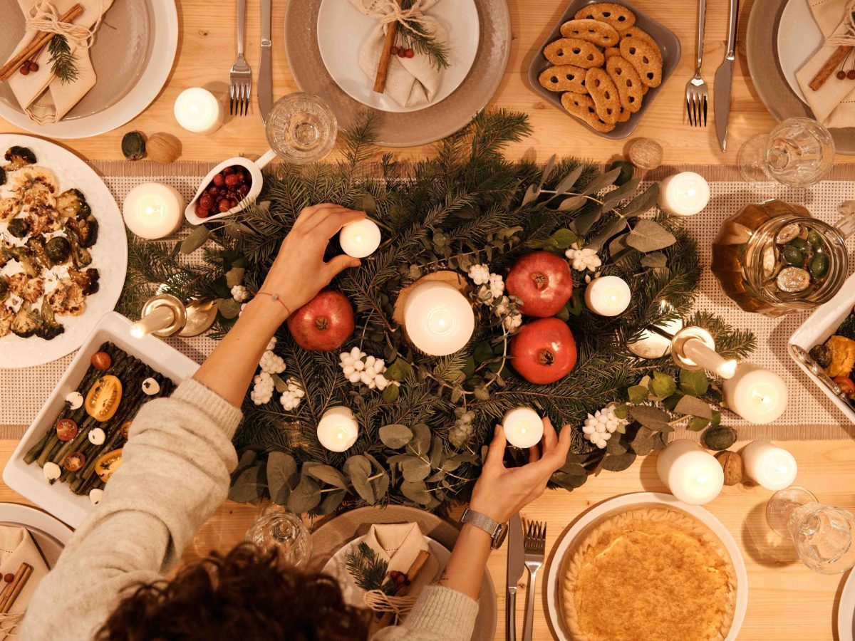A wooden table with a festive, winter center piece and many plates of food. Seen from above, there is a person arranging items in the center piece.