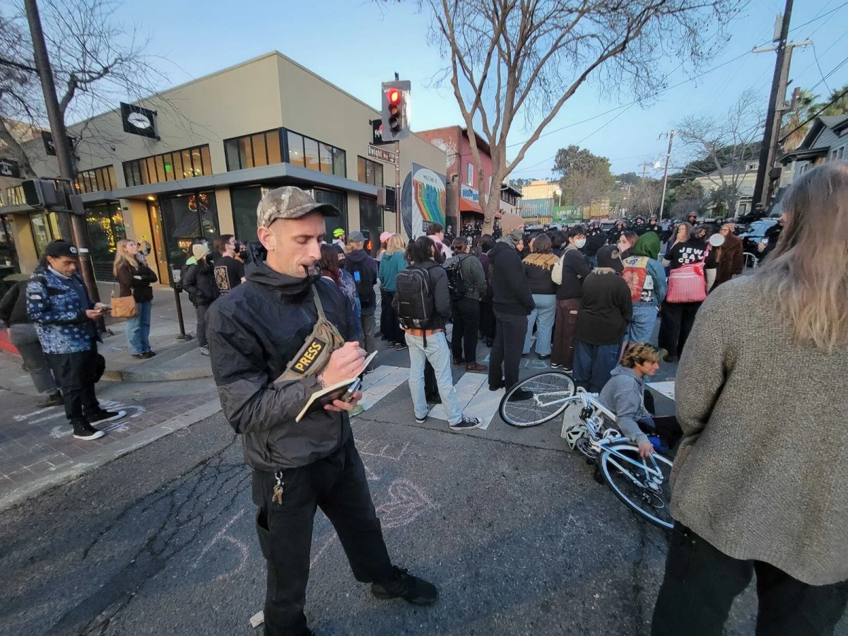A person with a press badge takes notes, while a pen dangles out of his mouth. He's a few steps away from a crown of dozens of people in an intersection, most bundled up in warm clothes.