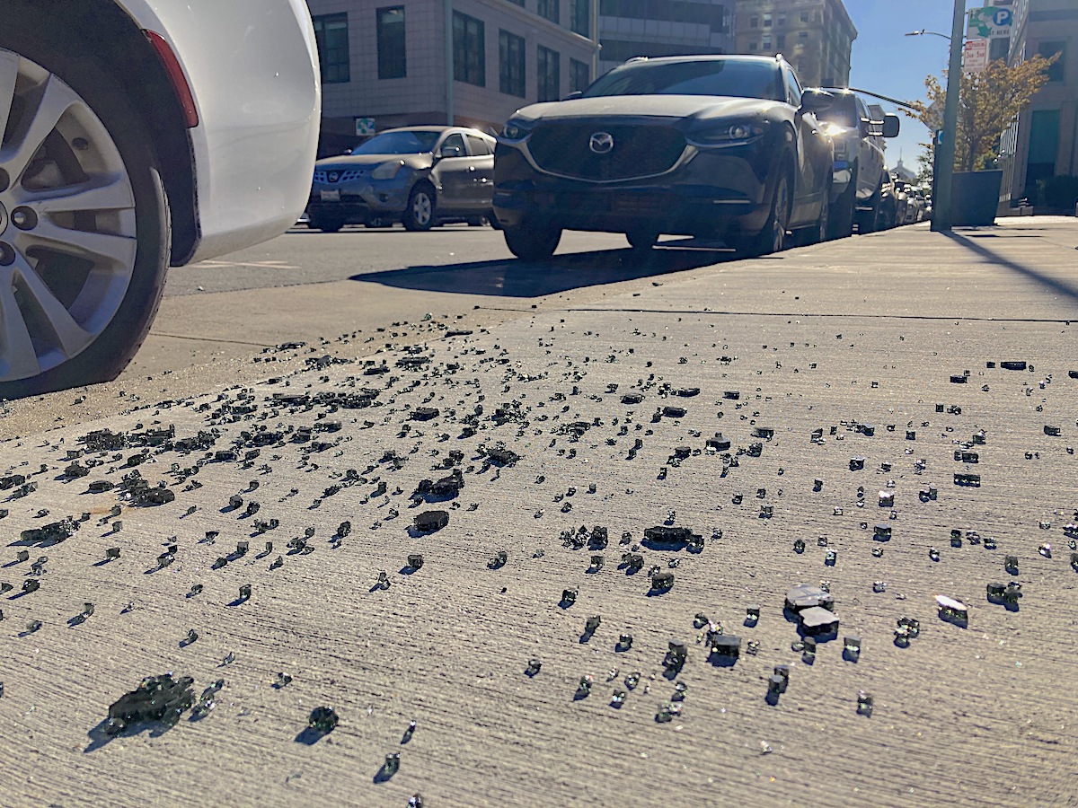 Bits of glass on the sidewalk with parked cars in the background.