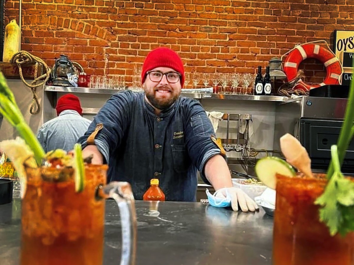 Oyster bar owner Branden Nichols stands behind a counter with a red knit hat on. Two micheladas in the foreground frame his torso.
