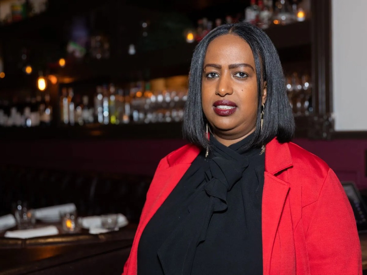Portrait of a Black woman in a black sweater and red coat standing inside a restaurant with a bar area in the background.