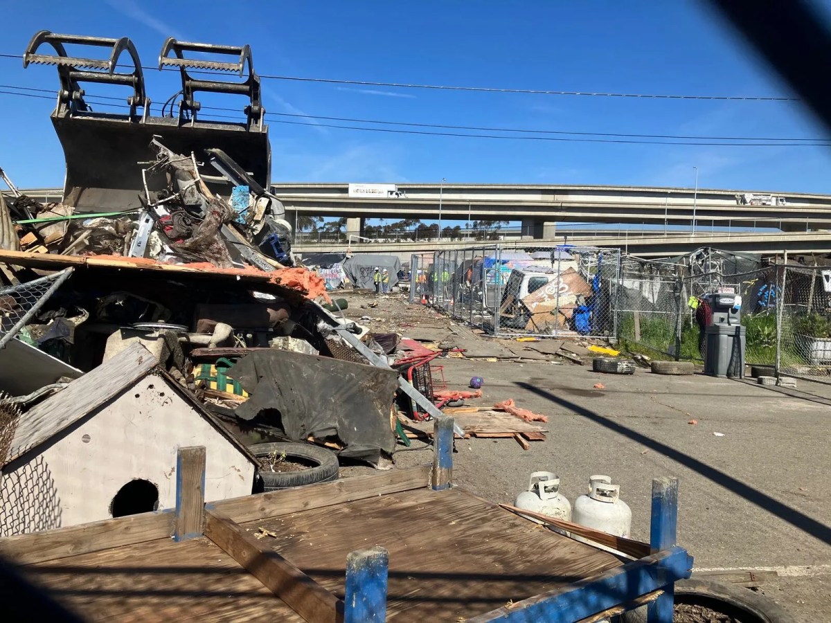 Teeth of a bulldozer preparing to clamp down on a pile of items at a homeless camp, with a freeway in the background.