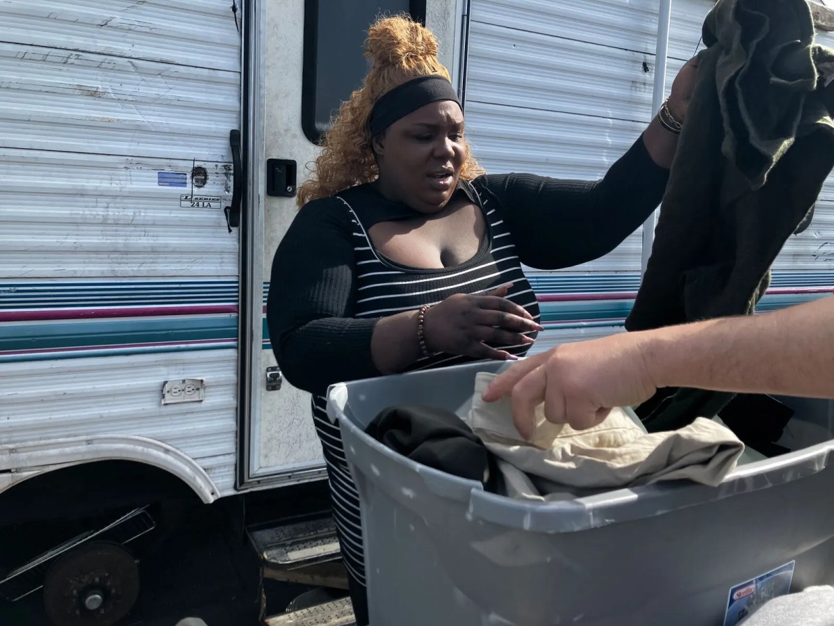 A woman standing in front of an RV holds up an item of clothing from a box filled with clothes.