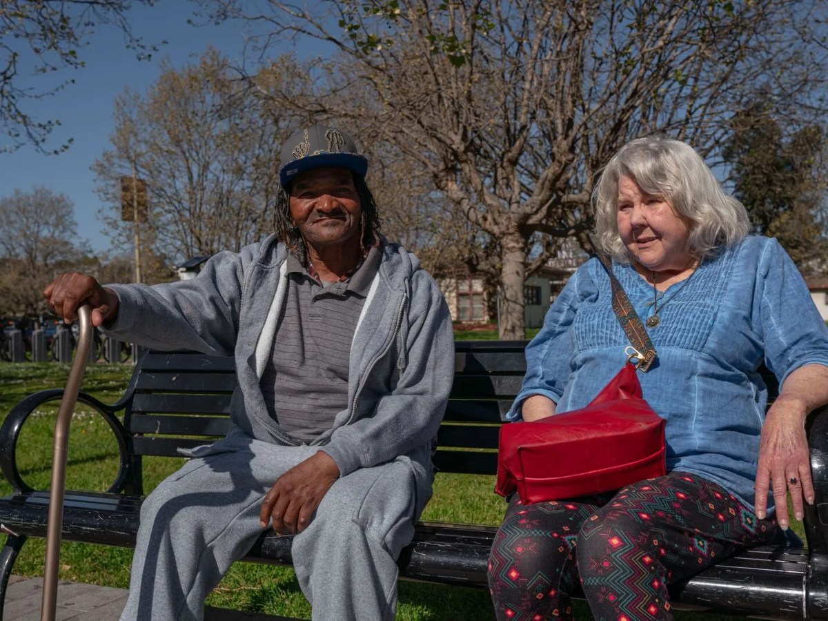 A middle-aged Black man with a cane and an older white woman sit together on a bench on a sunny day.