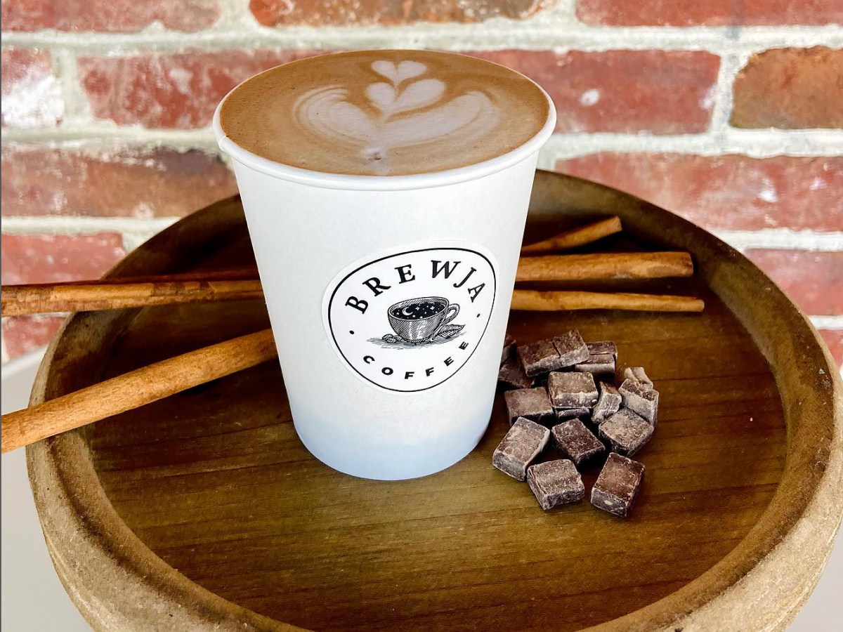 A latte from Brewja Coffee sits on a wooden table with some cinnamon sticks and chunks of chocolate.