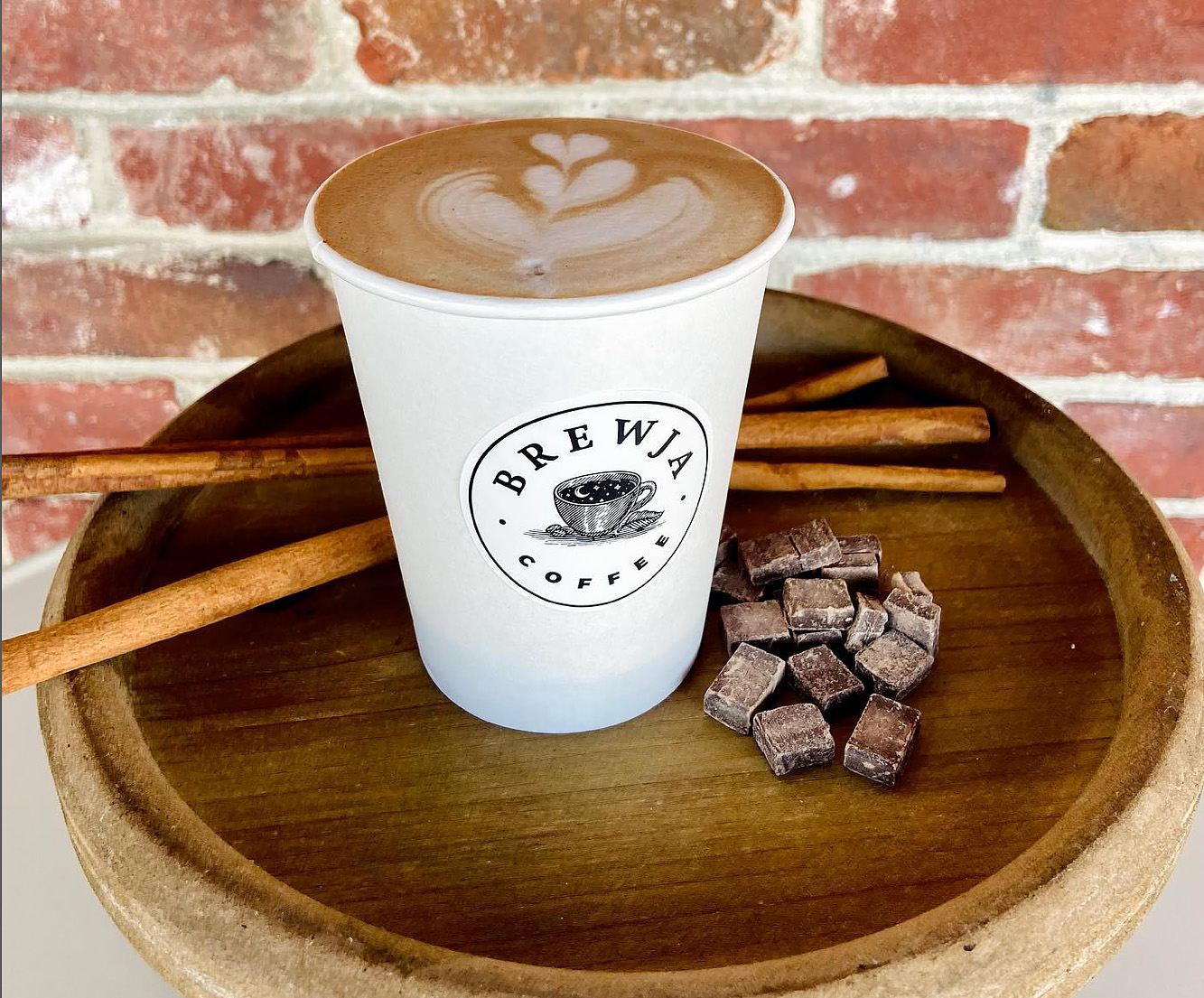 A latte from Brewja Coffee sits on a wooden table with some cinnamon sticks and chunks of chocolate.
