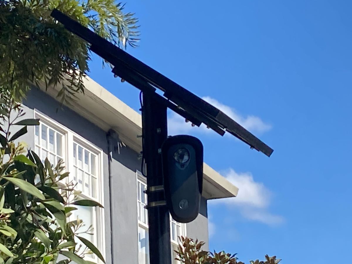 A black surveillance camera mounted on a pole underneath a solar panel on a steep street.