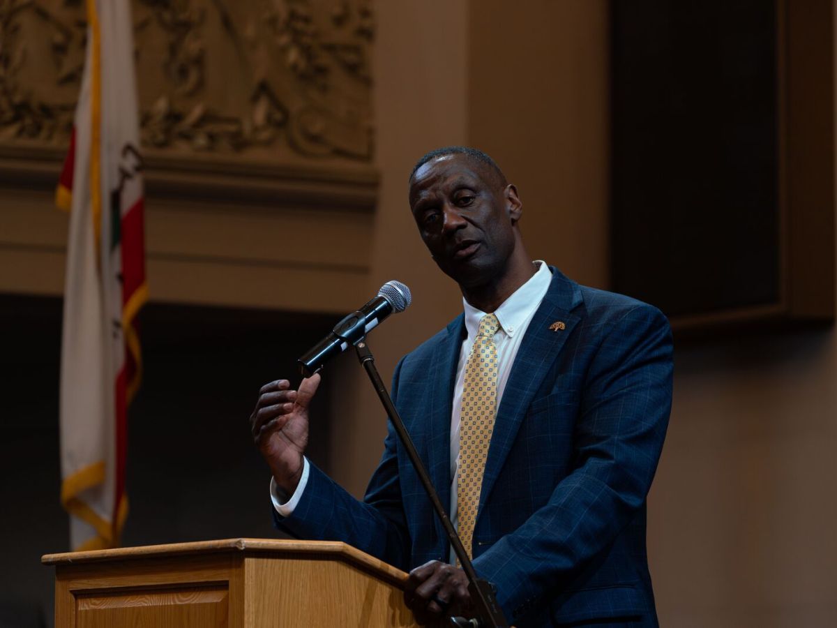 Man in blue suit speaking in front of a microphone and lectern in Oakland council chamber.