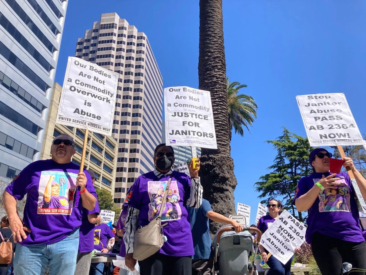 Three workers in purple t-shirts with union logos on them holding signs that read "our bodies are not a commodity," and "justice for janitors." Large office buildings loom in the background.