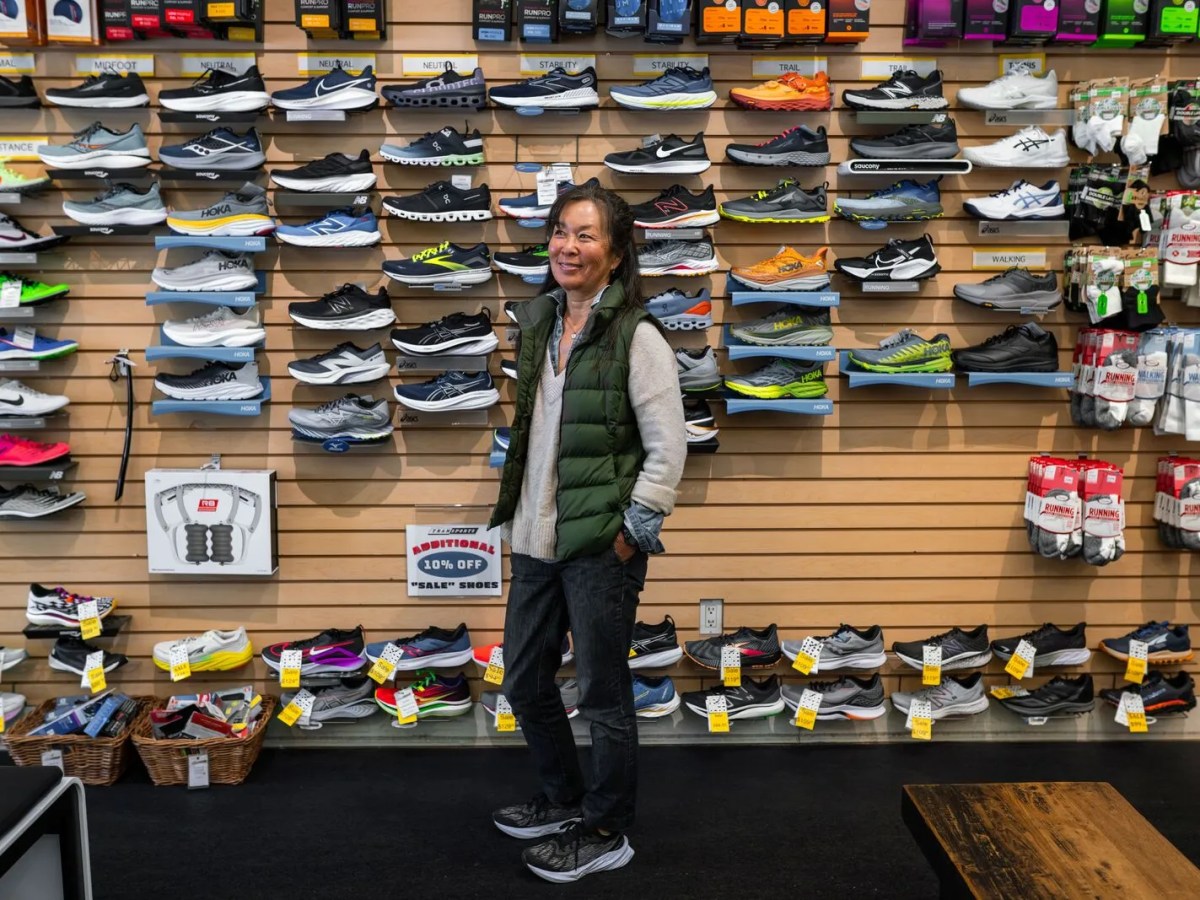 A woman wearing a green down vest, a gray sweater, blue jeans, and black athletic shoes smiles. Behind her is a wall filled with running and walking shoes of different colors, styles, and brands.