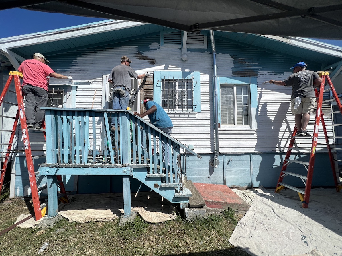 Four men paint a deteriorating blue house white.