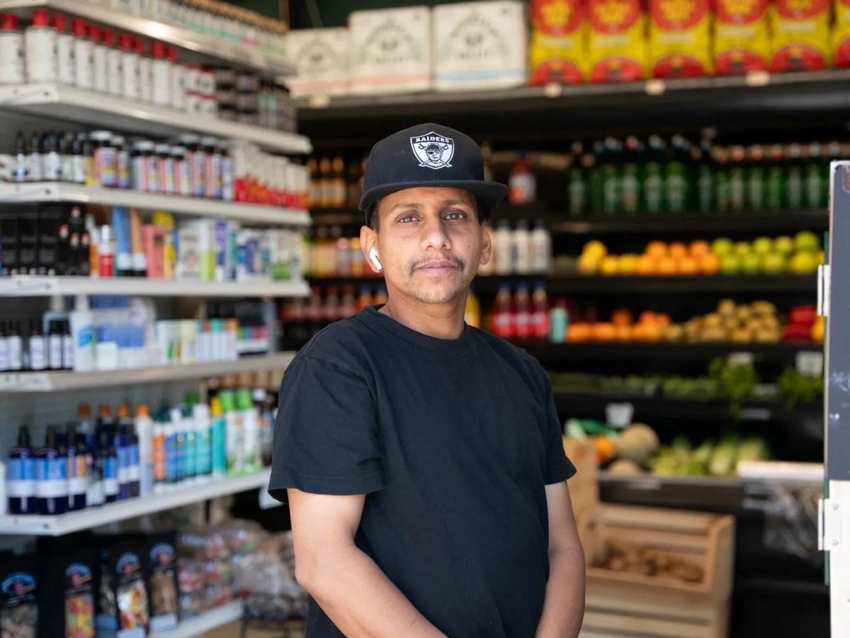 A man in a Raiders hat stands in a story with produce and other items behind him.